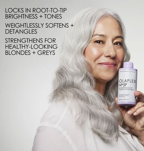 Smiling model with silver hair holds a bottle of Olaplex No. 5P Blonde Enhancer Toning Conditioner next to a caption of its benefits: Brightening, softening, detangling, strengthening