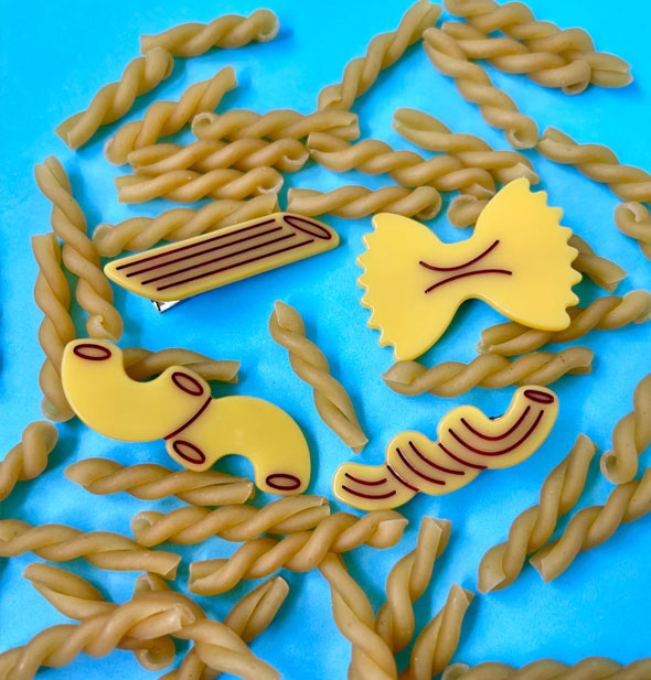 Set of four hair clips resembling pasta rest on top of gemelli scattered over a blue surface