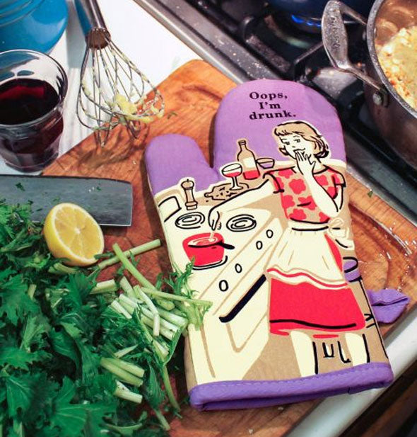 A colorful "Oops, I'm Drunk" oven mitt lays on a cutting board next to cut greens, lemon wedge, messy whisk, and wine glass