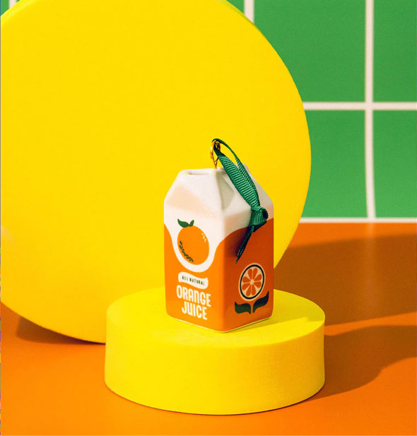 Orange juice carton ornament rests on a round yellow pedestal against a yellow, orange, and green backdrop