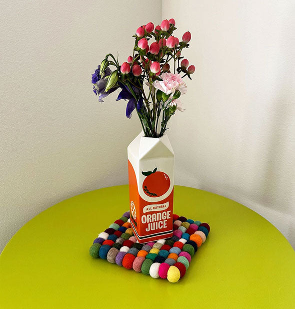 Orange Juice carton flower vase resting on a colorful woven coaster on a green tabletop holds a colorful bouquet