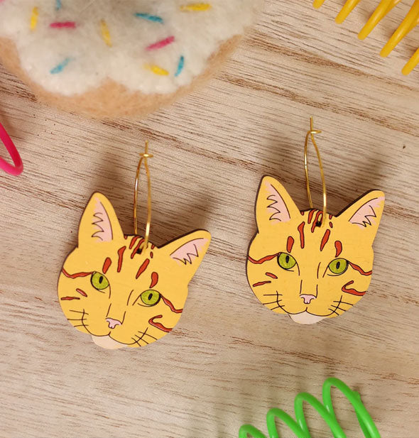 Pair of orange striped cat faces with green eyes hang from gold earring hoops and rest on a wooden surface with brightly colored coils and a plush toy