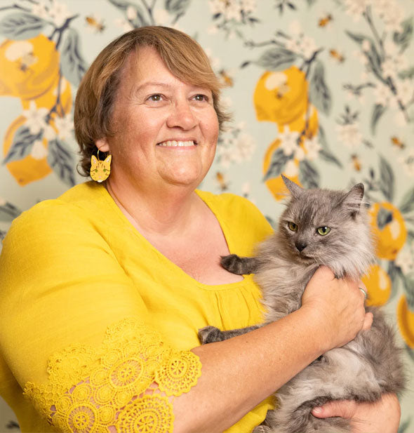 Smiling model wearing a pair of orange tabby cat face earrings holds a long-haired gray cat against a lemon wallpaper backdrop