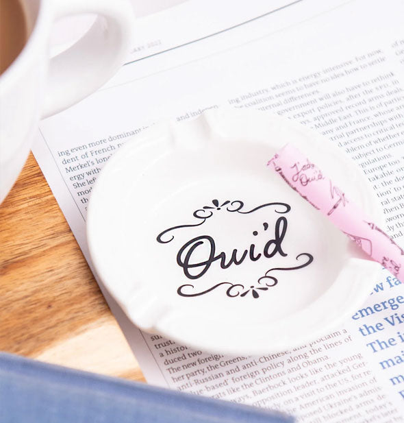 Round white ashtray resting on a newspaper with rolled pink cigarette proper says, "Oui'd" in the center in black script between two flourishes
