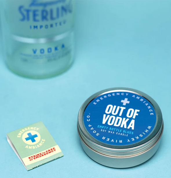 Out of Vodka Emergency Ambiance candle tin and matchbook rest on a blue surface with vodka bottle blurred slightly in the background