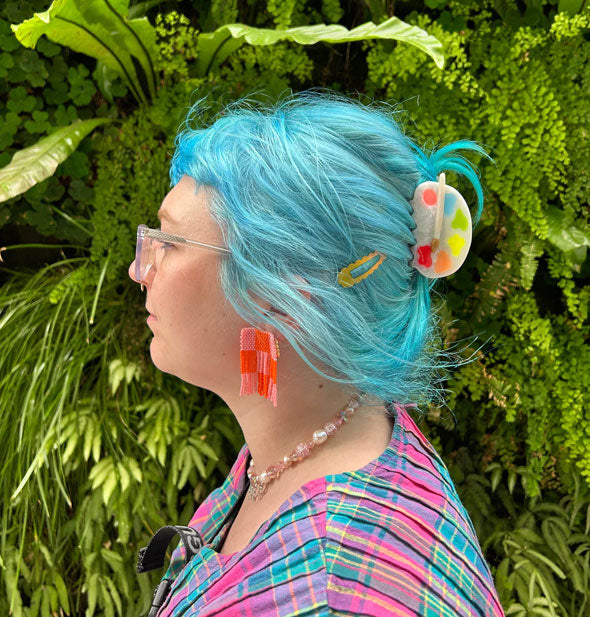 Model standing in front of a lush green botanical background wears a paint palette claw clip in a swept-back hairstyle