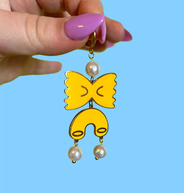 Model's hand holds a dangle earring by its gold hoop which features yellow bowtie and macaroni pasta charms accented by a white pearl at the top and two white pearls at the bottom
