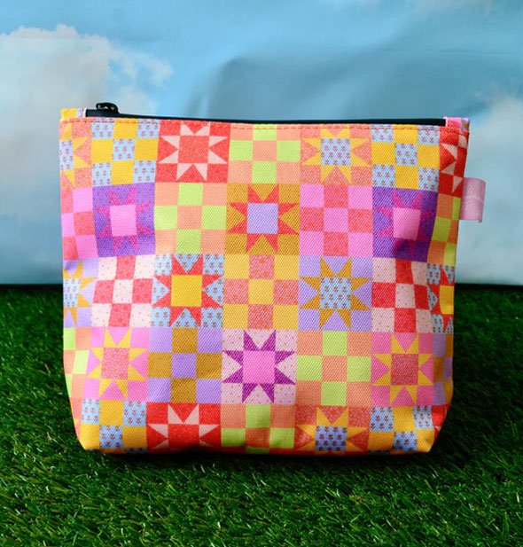 Colorful pouch with patchwork quilt design and a black zipper at top rests on green astroturf in front of a blue sky with clouds backdrop