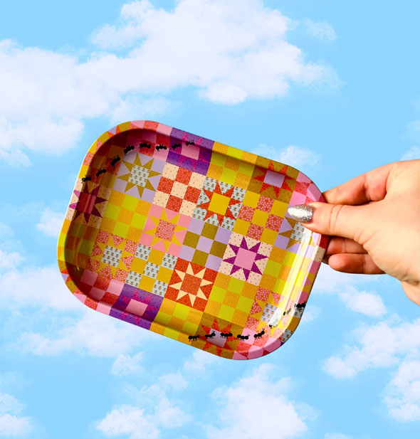 Model's hand holds a rectangular tray with rounded corners and an all-over colorful patchwork design up in front of a blue sky with clouds backdrop