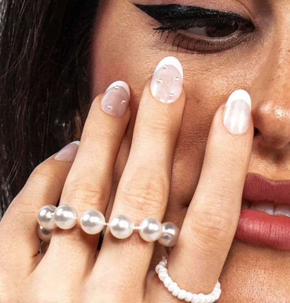 Model with pearl-accented French tip press on nails and accessories lightly touches face with fingertips