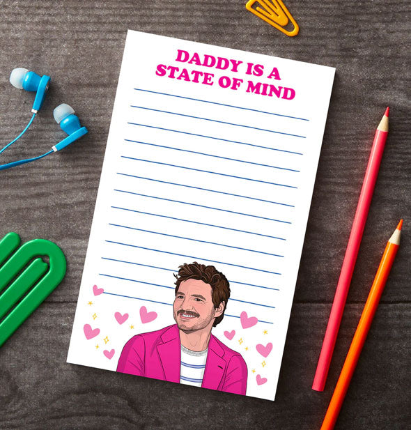 Daddy Is a State of Mind Pedto Pascal notepad rests on a wooden surface with green and yellow paper clips, blue earbuds, and red and orange colored pencils