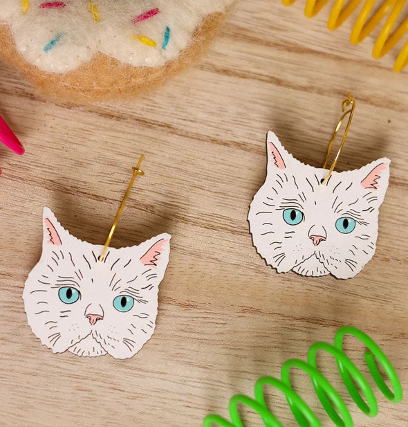 White Siamese cat faces with blue eyes hang from gold earring hoops on a wooden surface scattered with brightly colored coils and a plush toy