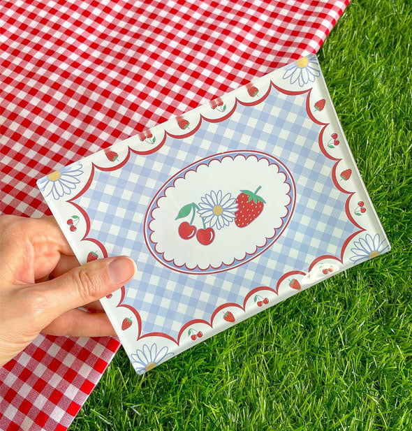 Model's hand holds a rectangular tray with designs of cherries, strawberries, and daisies accented by a red border and central blue gingham in front of a backdrop of green astroturf and a red gingham tablecloth