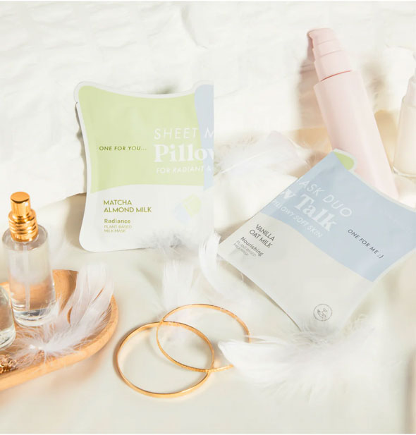 Matcha Almond Milk and Vanilla Oat Milk Pillow Talk sheet mask packettes rest on a plush white surface scattered with feathers, accessories, and toiletry bottles