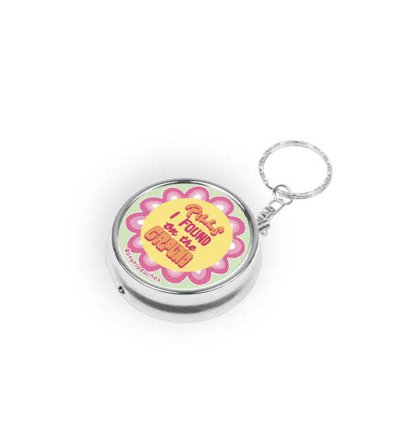 Round silver pill case on keychain attachment says, "Pills I found on the ground" in orange and pink lettering inside a yellow circle with pink petal-like border
