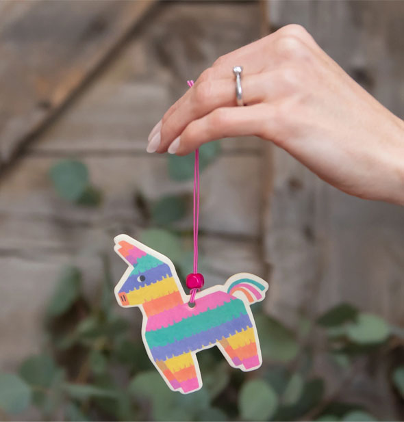 Model's hand holds a colorful piñata car air freshener by its pink string in front of a botanical and wood backdrop
