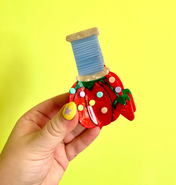 Model's hand holds a hair clip designed to resemble a spool of blue thread atop a tomato pincushion with multicolored pin heads sticking out of it