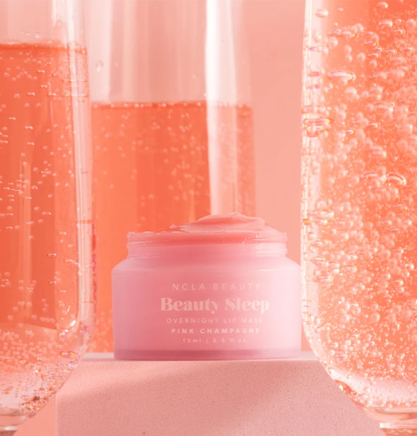 Opened pot of Pink Champagne NCLA Beauty brand Beauty Sleep Overnight Lip Mask staged with flutes of bubbling pink champagne