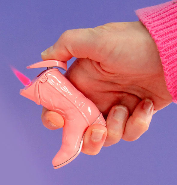 Model's hand holds a lit pink cowgirl boot cigarette lighter with a pink flame against a purple backdrop