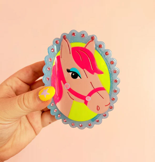 Model's hand holds a hair clip with oval cameo-style design of a pink pony on a yellow background inside a blue scalloped border accented with pink rhinestones