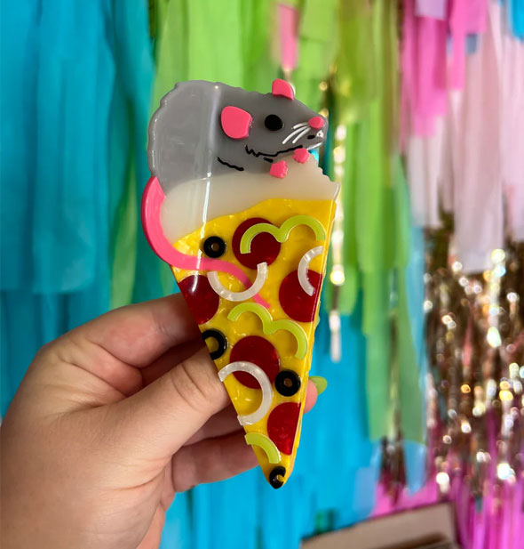 Model's hand holds a hair clip designed as a piece of supreme pizza with a rat on top in front of a colorful streamer backdrop