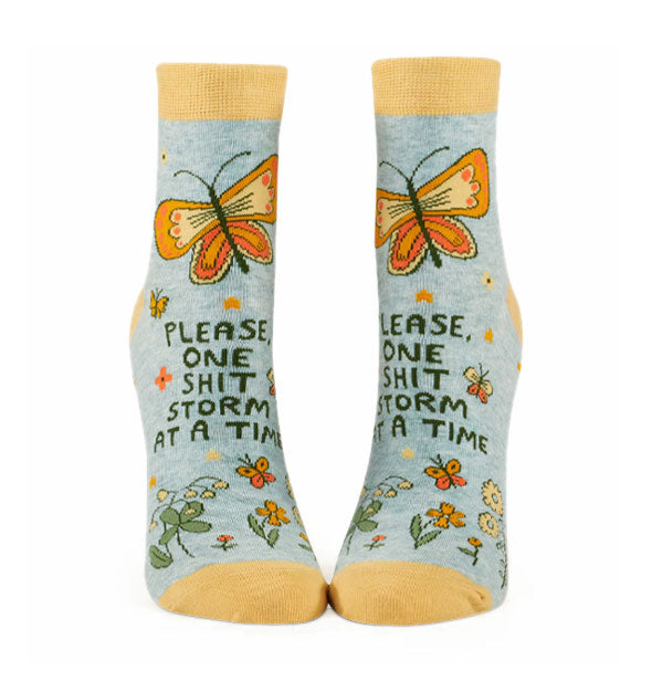 Pair of gray ankle socks with yellow accents feature colorful floral and butterfly designs around the words, "Please, one shit storm at a time"