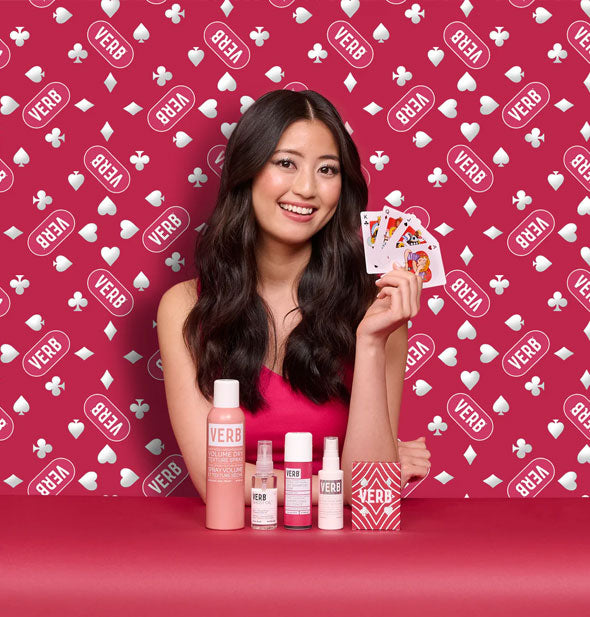 Smiling model holding a hand of playing cards against a red and white patterned background poses with the products in Verb's Poker Face, Perfect Hair Best-Selling Stylers Kit