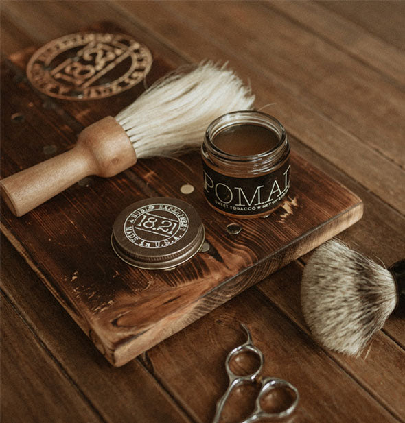 An opened jar of hair styling Pomade rests on a wooden board with men's grooming tools