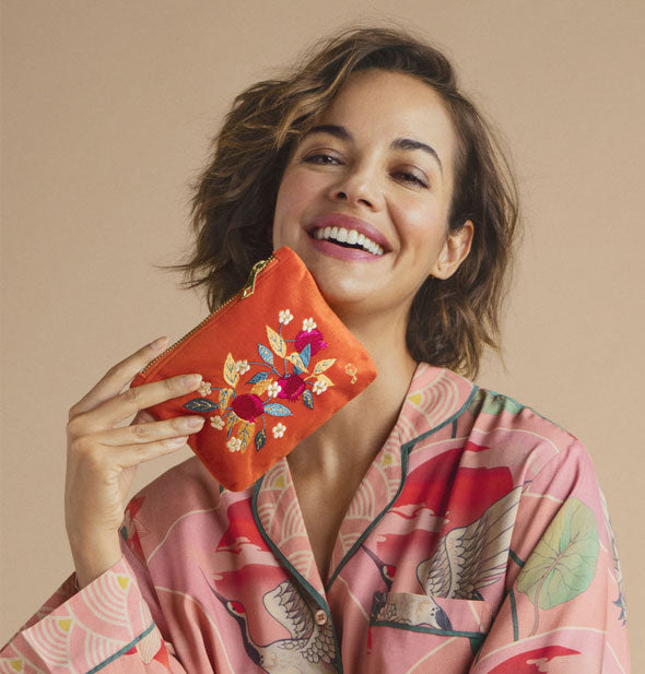 Smiling model holds an orange velvet embroidered pomegranate pouch to chin