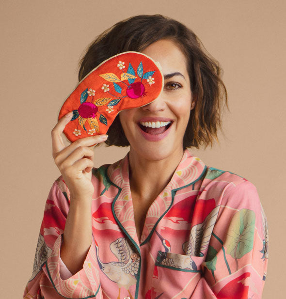 Smiling model holds an orange velvet Pomegranate Sleep Mask playfully over one eye