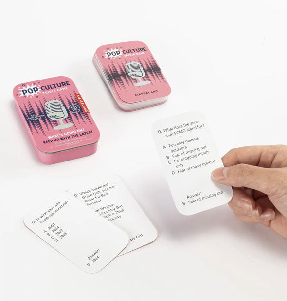 Model's hand holds a card from the Pop Culture Trivia Game; two other cards rest nearby along with the tin and deck beyond