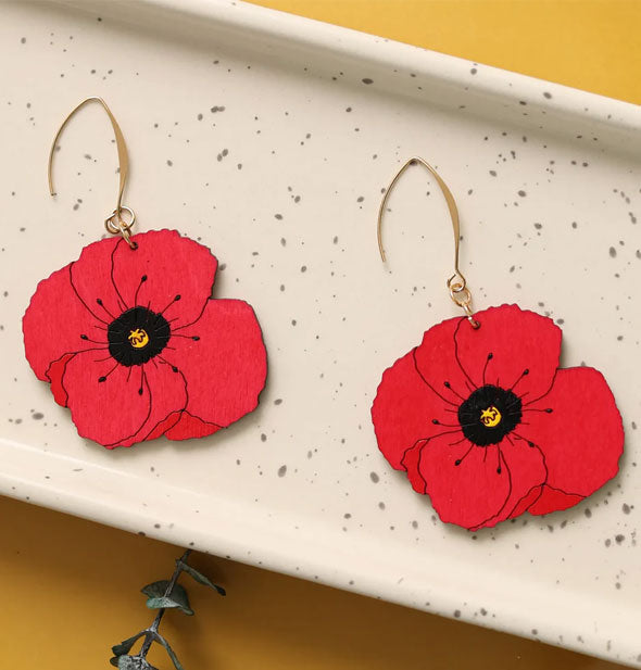 Pair of red poppies with black and yellow centers on gold earring hooks rest in a white speckled dish on a yellow surface
