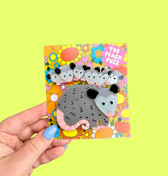 Model's hand holds a colorful floral-patterned product card with possum hair clips attached: one larger "mama" possum and one with a lineup of six "babies"