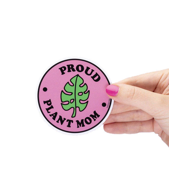 Model's hand holds a round sticker with green monstera leaf illustration and the words, "Proud Plant Mom" on a pink background with white and black border