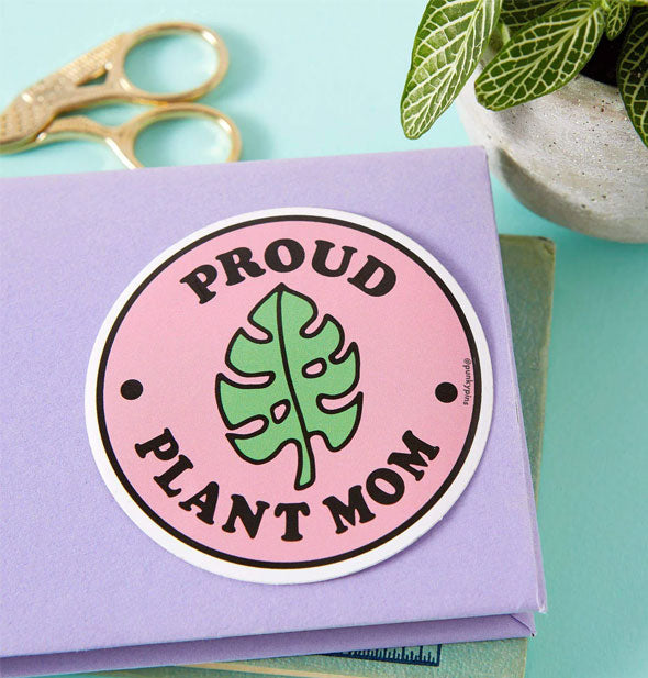 Proud Plant Mom sticker on a purple notebook cover staged with houseplant and gold shears