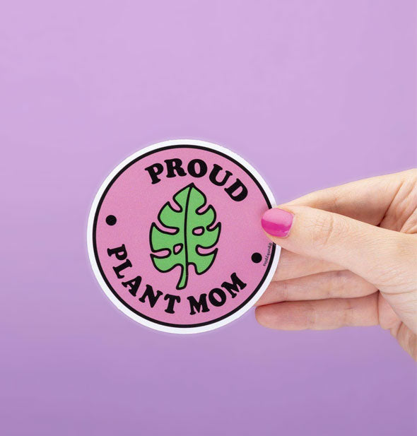 Model's hand holds a round sticker with green monstera leaf illustration and the words, "Proud Plant Mom" on a pink background with white and black border
