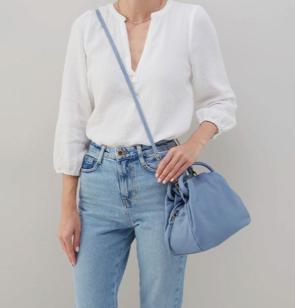 Model wearing jeans and a white shirt wears a pale blue leather satchel bag in a crossbody fashion with an elongated strap