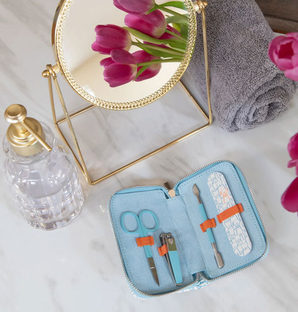 Blue manicure pouch lays open on a marble countertop with round gold mirror, crystal soap dispenser, pink tulips, and gray towel