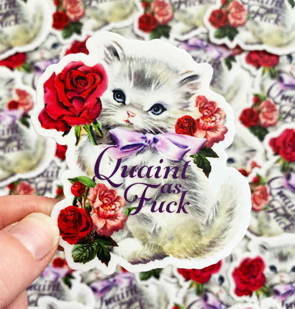 Model's hand holds a Quaint as Fuck kitten sticker in front of a pile of others like it in the backgrounf