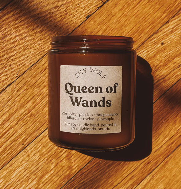 Shy Wolf 8 ounce amber glass jar soy candle on wood floor with label that says, "Queen of Wands: Creativity, passion, independence, hibiscus, melon, pineapple"