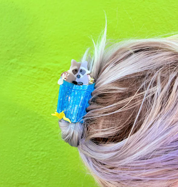 Model's hair is styled with a claw clip designed as a raccoon emerging from a blue trash can surrounded by scraps