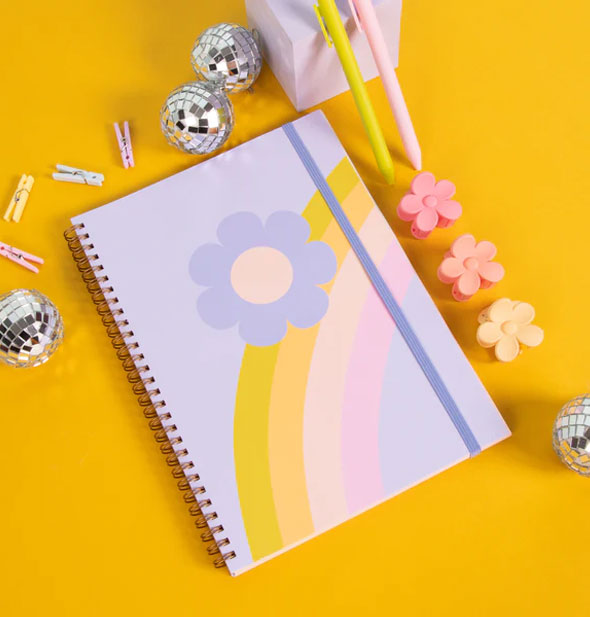 Rainbow Flower Spiral Notebook is staged with mini clothespins, flower clips, pens, and mini disco balls on a mustard yellow surface