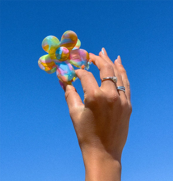 Model's hand holds a rainbow swirl flower-shaped hair claw clip up in front of a clear blue sky