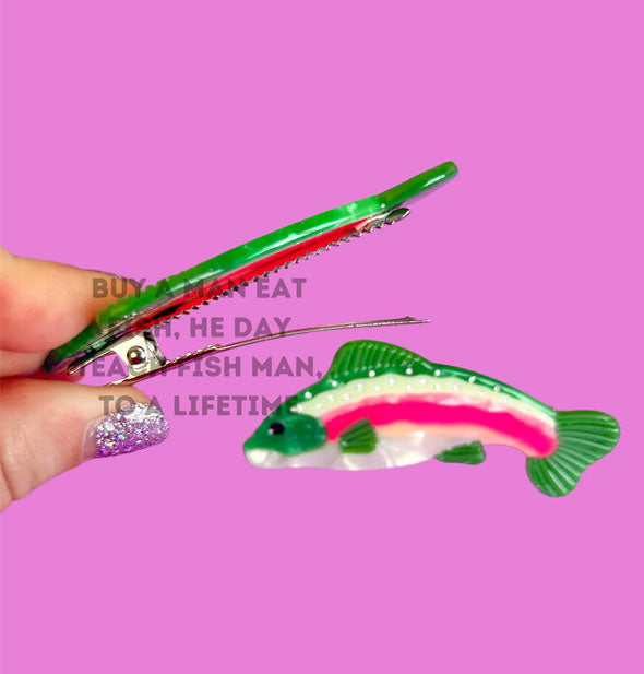 Model/s hand holds a rainbow trout hair clip alligator-style clip open as another like it rests in the background; a watermark overtop the image reads, "Buy a man eat fish, he day teach fish man, to a lifetime"