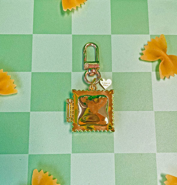 Square gold ravioli locket with hinge and attached clasp hardware rests on a checkered green surface scattered with farfalle