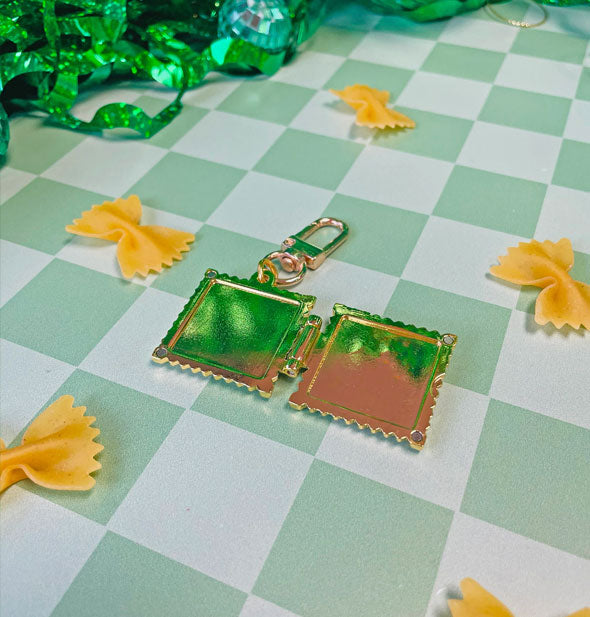 Opened square gold ravioli locket keychain rests on a checkered green surface scattered with farfalle and shiny green streamers and disco balls