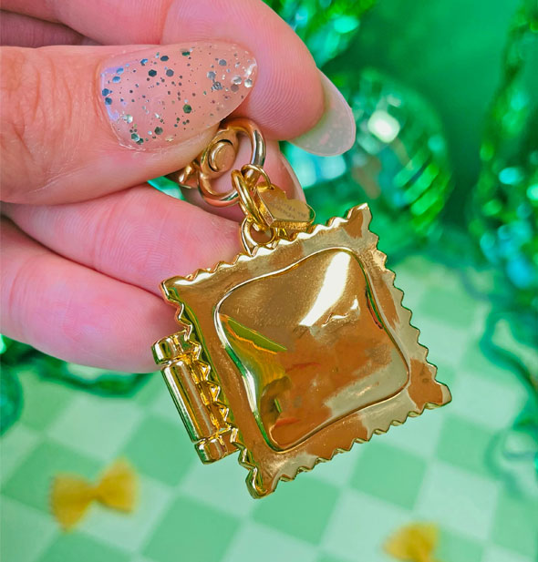 Model's hand holds a gold ravioli locket keychain by its clasp against a green backdrop