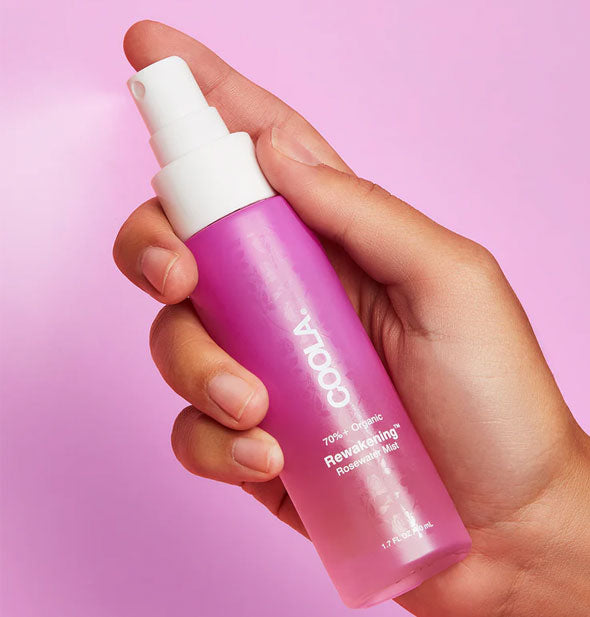 Model's hand dispenses a fine spray from a bottle of Coola Rewakening Rosewater Mist against a pink background