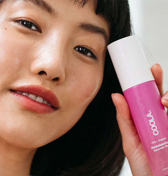 Model with dewy complexion holds a bottle of Coola Rewakening Rosewater Mist