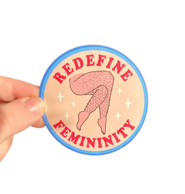 Model's hand holds a round sticker with blue border that features an illustration of crossed stubbly legs accented with white stars and framed by the words, "Redefine femininity" at top and bottom in red lettering